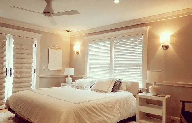 A cozy bedroom with a large bed, white bedding, and two bedside tables with lamps. The room has soft lighting, a ceiling fan, and closed blinds on the windows.