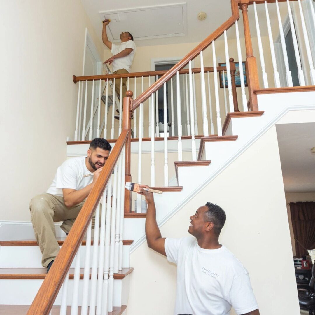 Three men in white shirts work on a staircase. One stands at the bottom, one kneels on the steps, and another adjusts a ceiling fixture at the top.