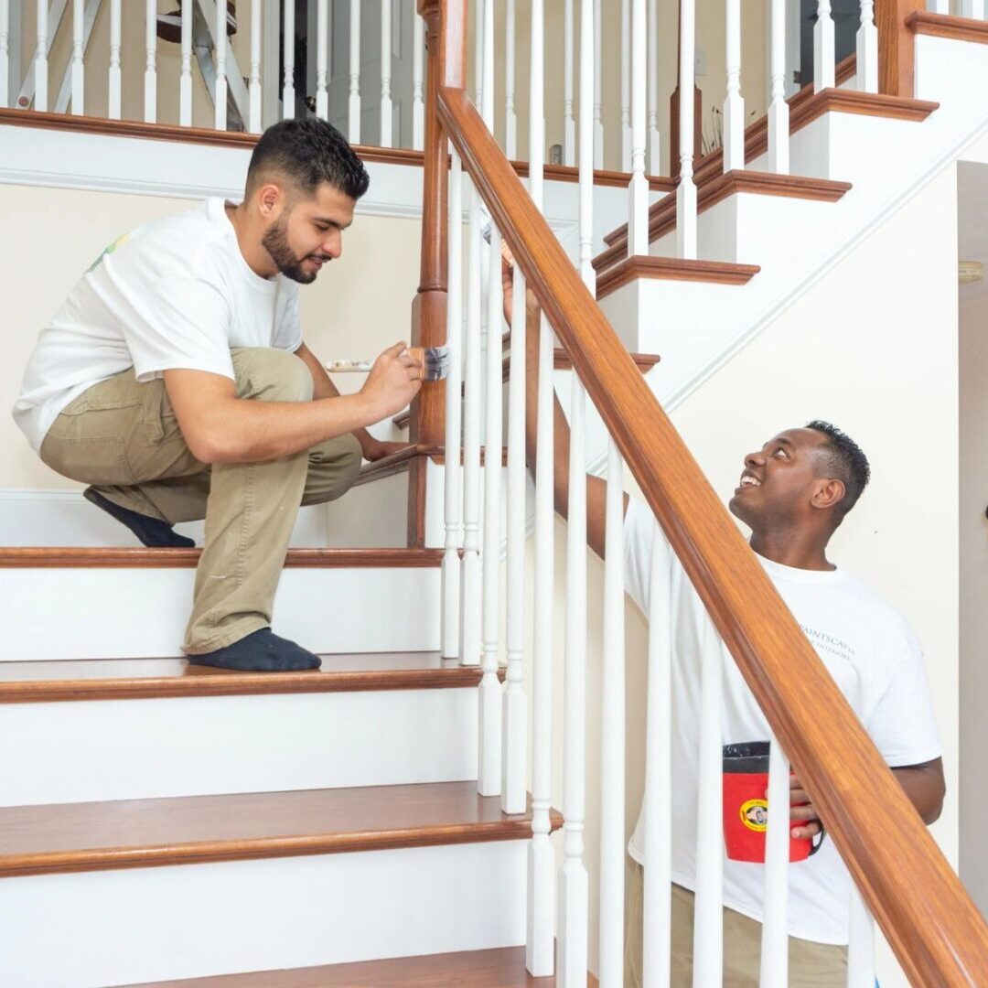 Two men are painting a staircase. One is kneeling on the steps, holding a brush, while the other stands, holding a paint container. A sign on the wall says "Home.