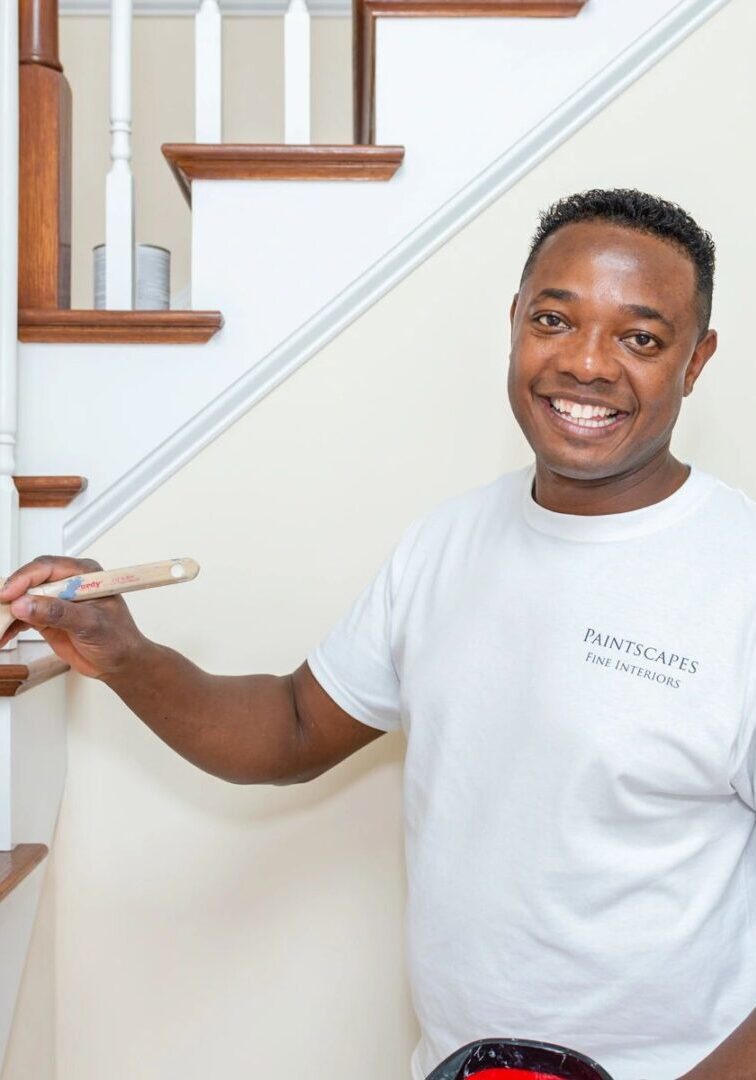 A person holding a paintbrush smiles while painting a staircase railing, with another person sitting on the stairs in the background.